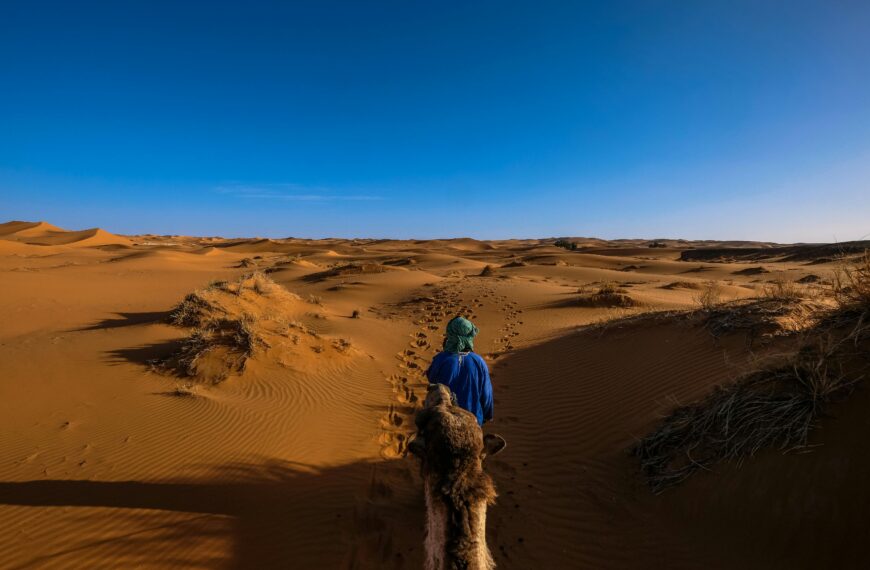Person riding a camel through the dunes of Ksar Tanamouste, Morocco under a clear blue sky.